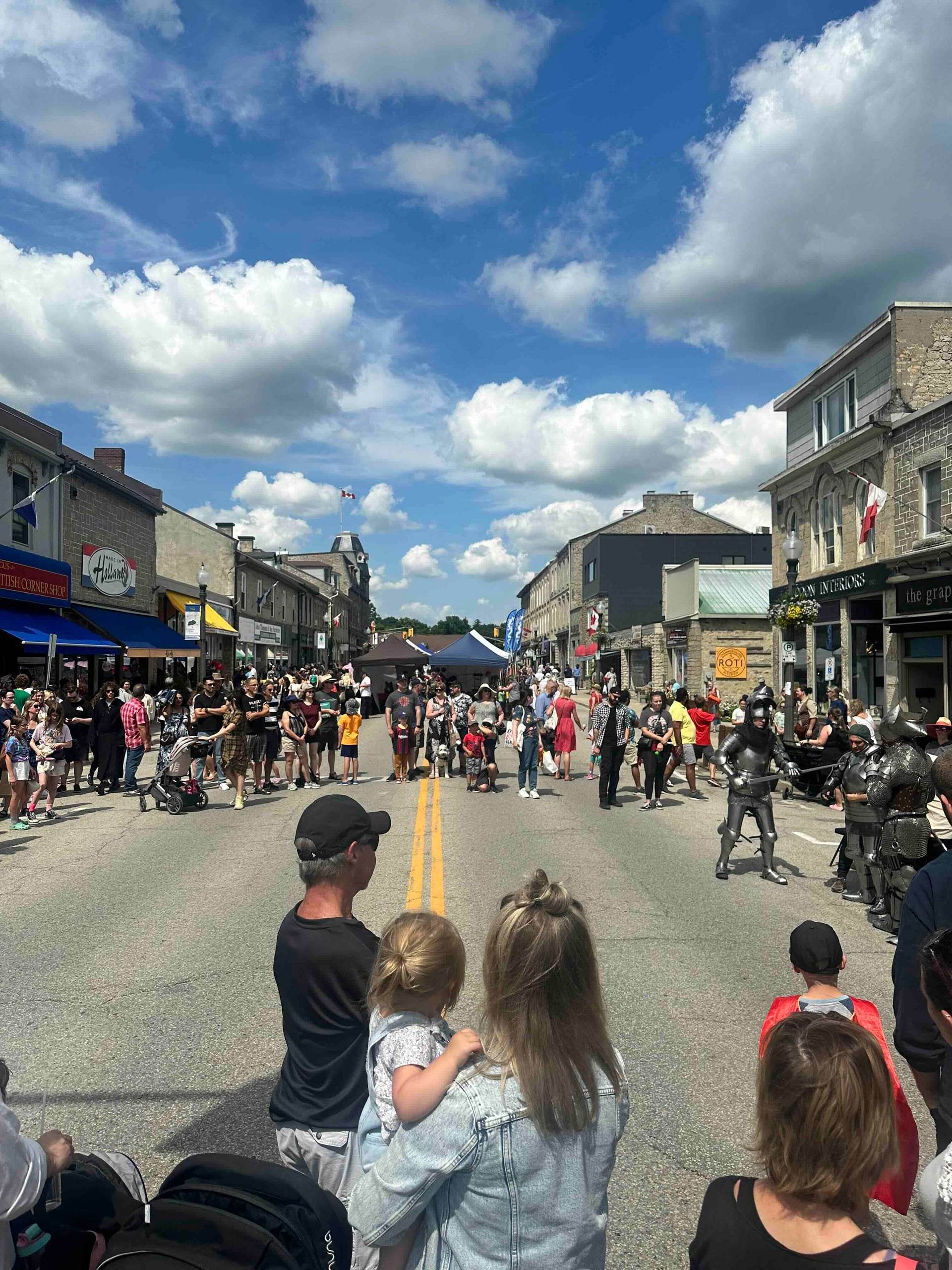 A summer street festival on the main street in downtown Fergus, with stone storefronts and performers in armour.