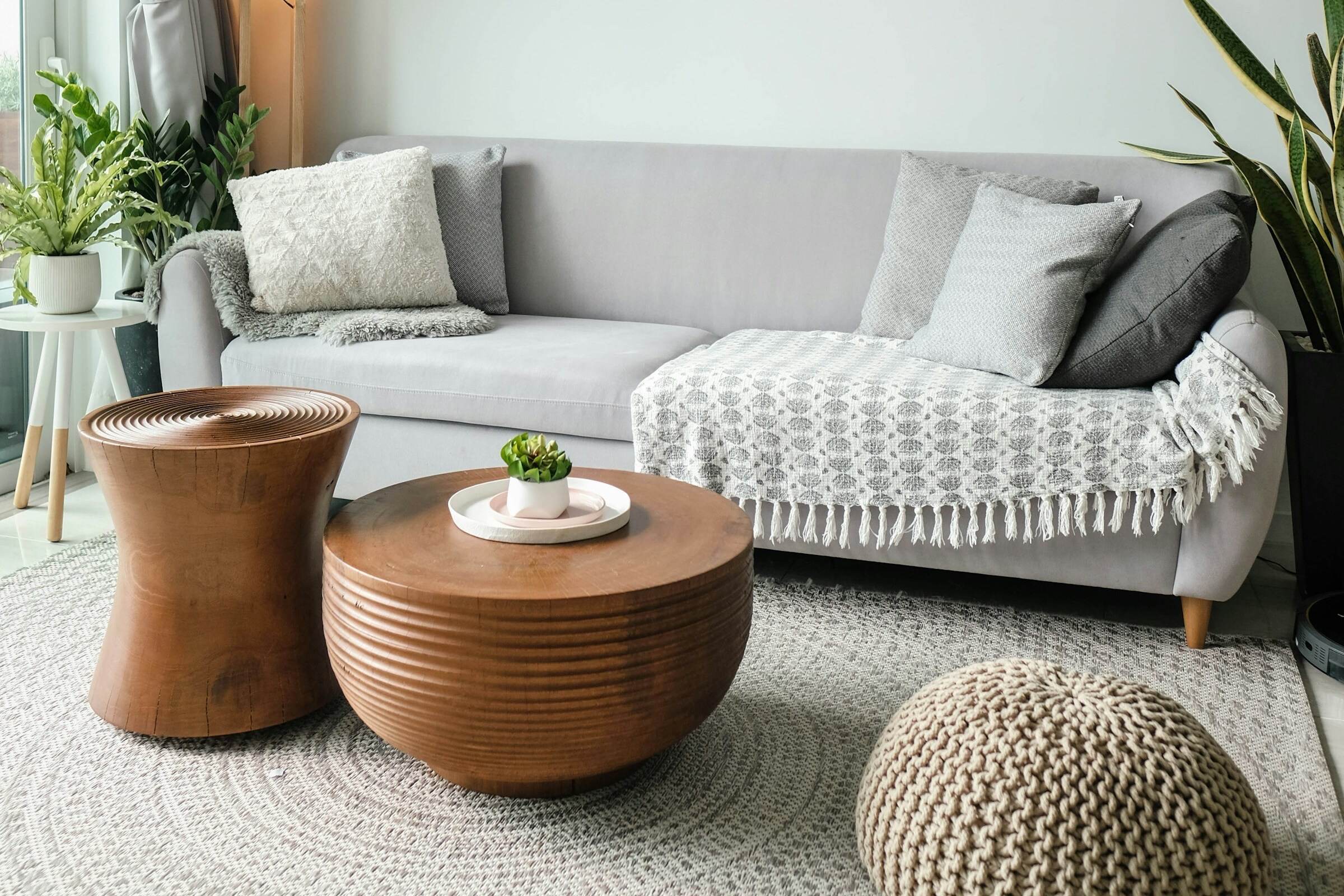 A bright living room with a grey sofa, throw blanket, round wooden tables and a woven rug.