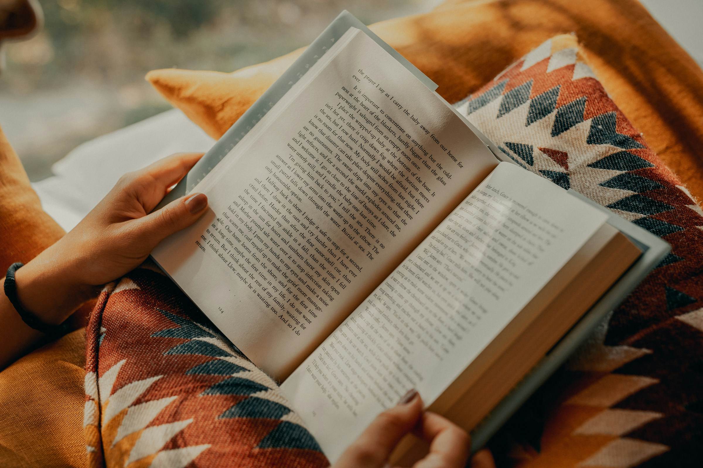 A person reading an open book on a blanket with a patterned cushion.