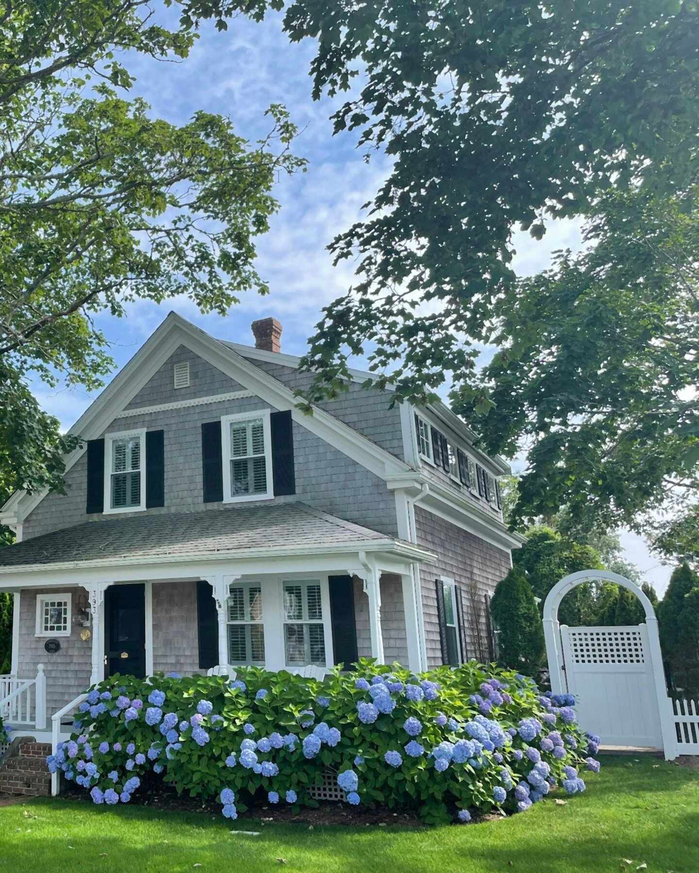A grey-shingled two-storey house with blue hydrangeas along the front and a white garden gate.