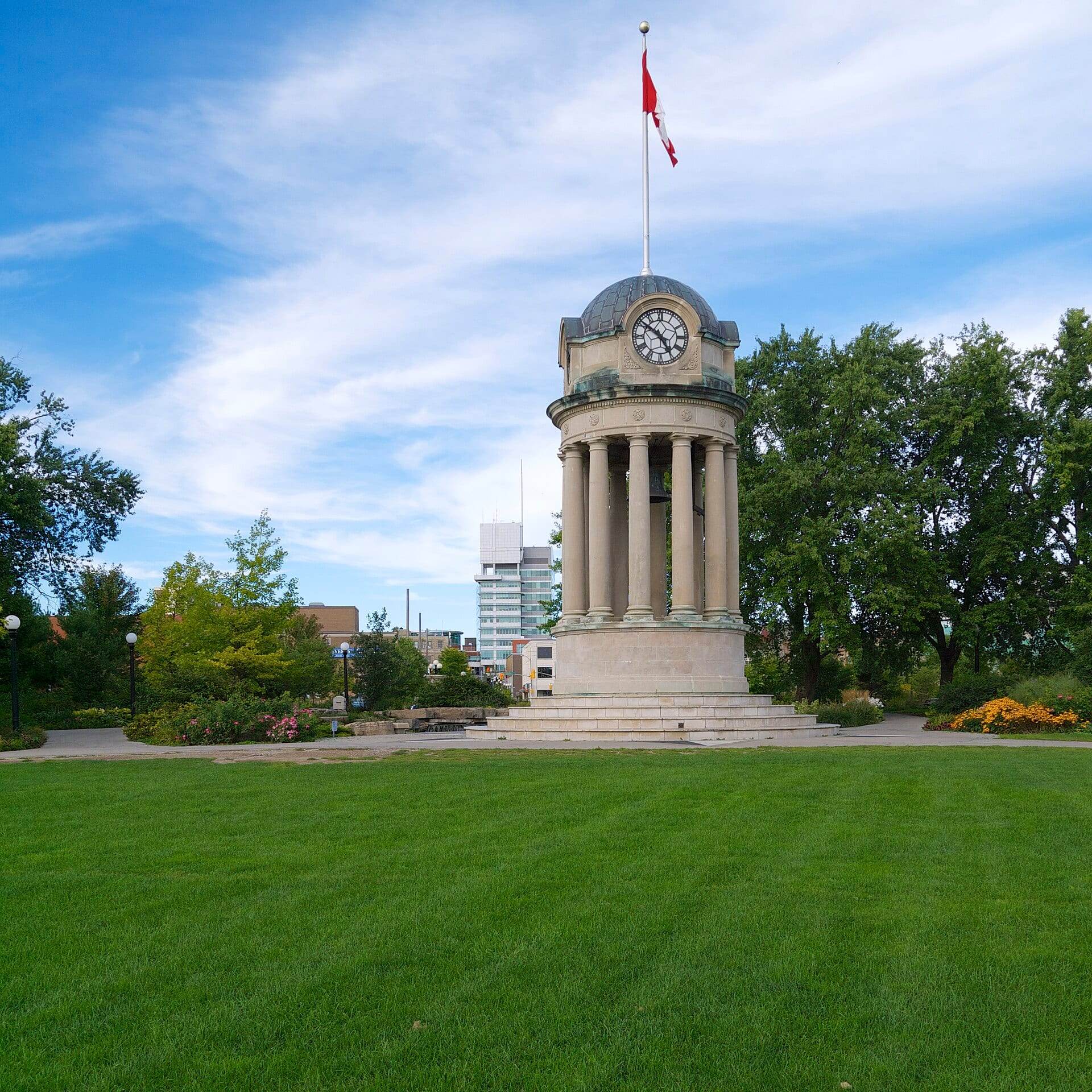 The clock tower in Kitchener's Victoria Park on a sunny day, with downtown buildings behind the trees.
