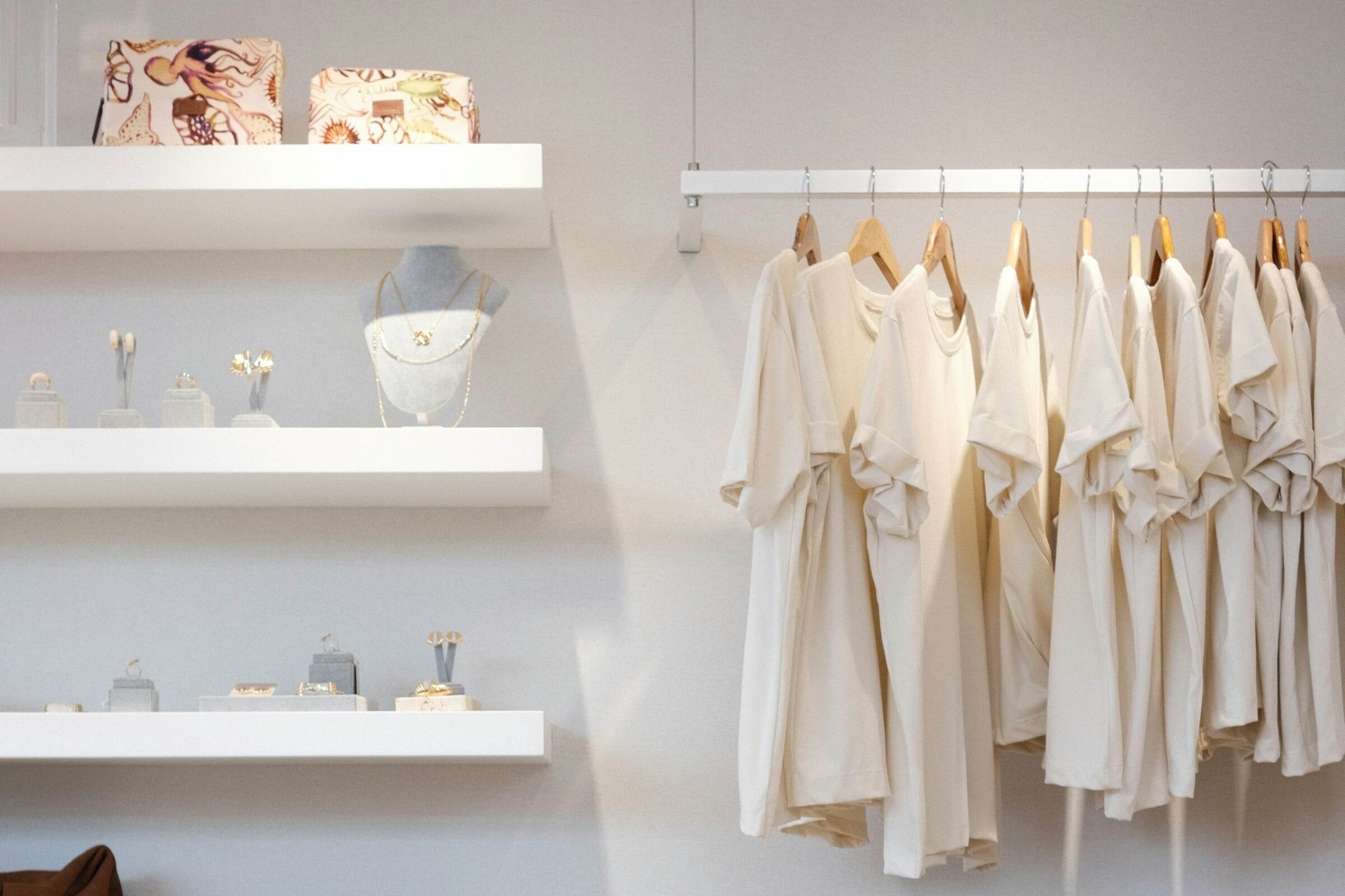 A bright boutique with garments on a rail and goods on white wall shelves.